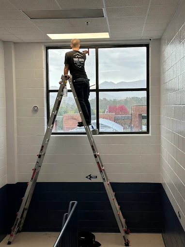 True South installer on a ladder applying 3M safety film to a school hallway window with mountains visible outside